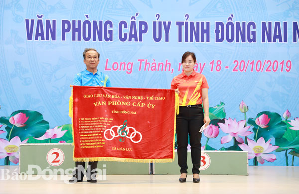 Chánh văn phòng Huyện ủy Long Thành Huỳnh Minh Thảo (trái) trao cờ luân lưu đăng cai giao lưu cho Phó chánh Văn phòng  Tỉnh ủy Nguyễn Thị Hồng Trang – đại diện cho đơn vị đăng cai tổ chức giao lưu văn nghệ - thể thao truyền thống các ban Đảng và văn phòng cấp ủy lần tới vào năm 2020.