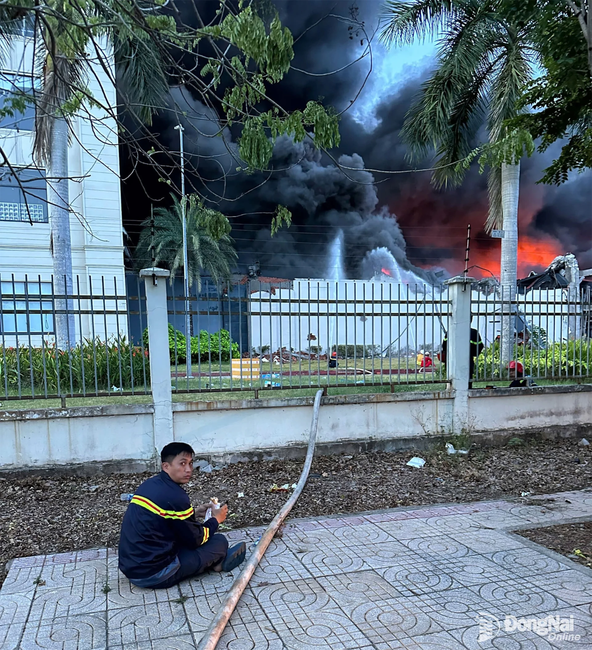 Một chiến sĩ tranh thủ ăn vội bên ngoài hiện trường, phía sau là khói lửa từ vụ cháy Công ty TNHH Uniwin Việt Nam, ngày 10/5/2026.