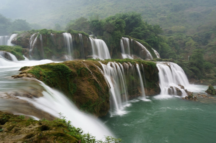 The waterfall is separated into three layer-falls by rocks and trees. Large volumes of water pouring down to lime stone steps form a curtain of white spray.